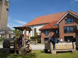 Außenansicht der Kinderkrippe St. Josef. Modernes Gebäude, davor ein Spielplatz mit spielenden Kindern. Im Hintergrund die St. Vitus Kirche und auf der linken Seite des Bildes der Kirchturm.